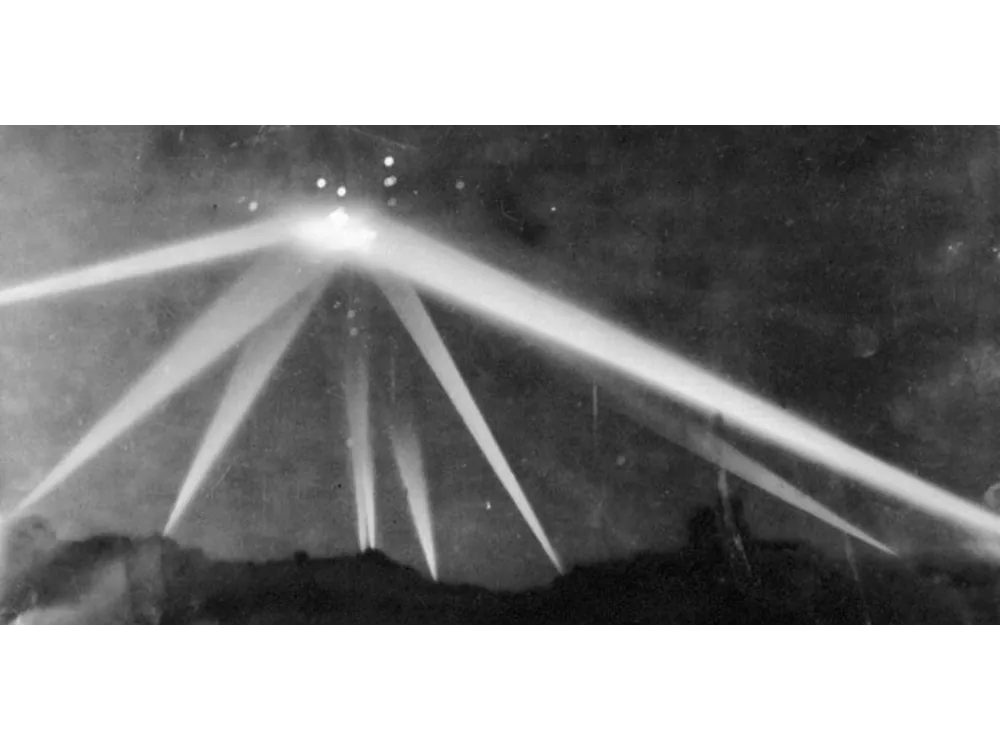 Night photograph of searchlight beams converging on a point in the sky above the city.