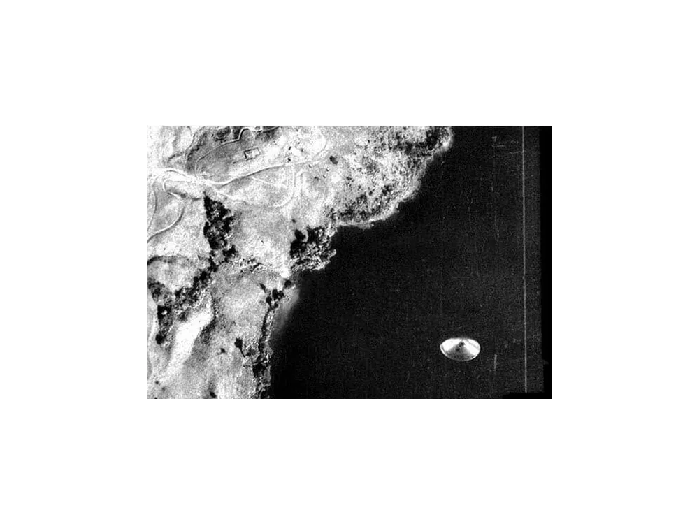 Aerial survey photograph of coastline and terrain, with a bright disc-shaped object at the lower right.