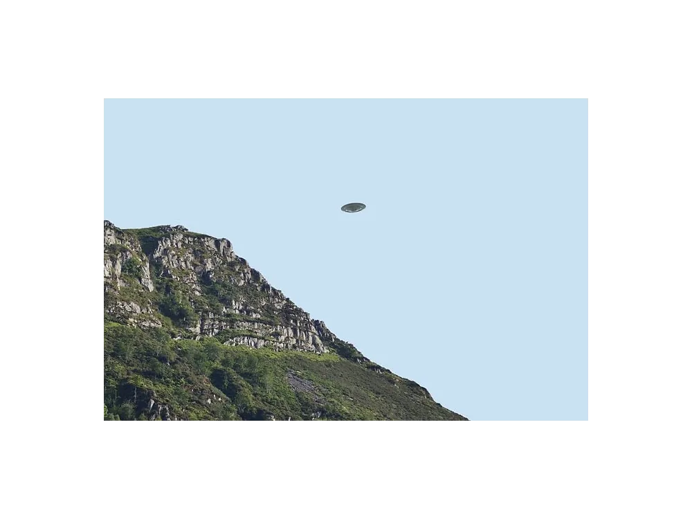 Colour photograph of a pale disc in a clear sky beyond a steep green headland.