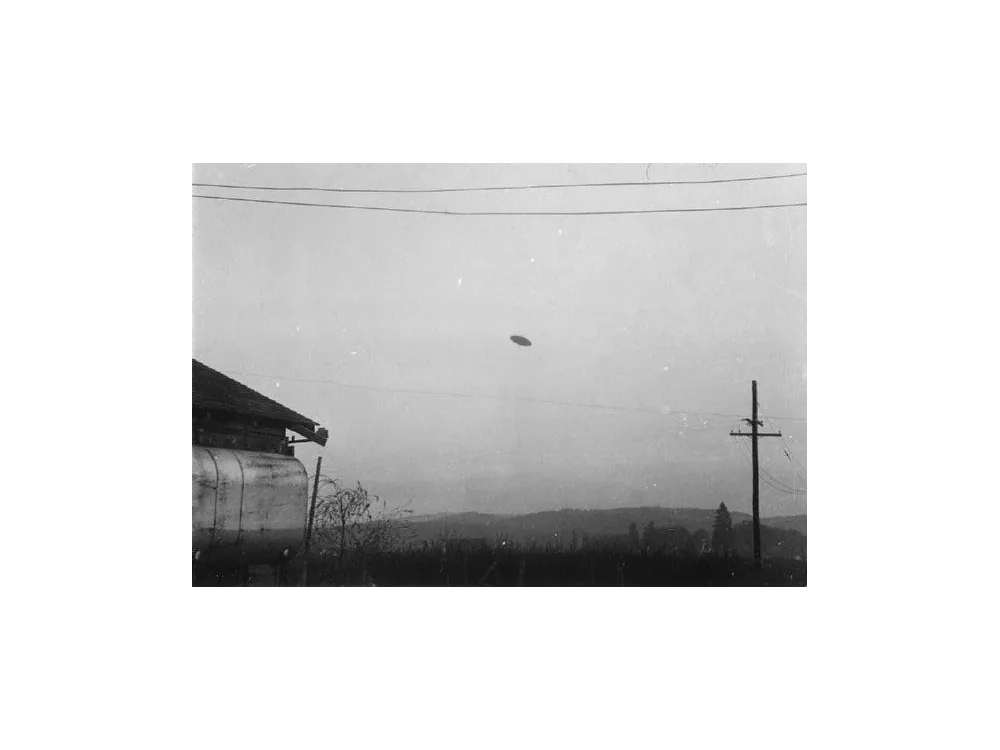 Black and white photograph of a disc above a town, with power lines and a barn roof in the foreground.